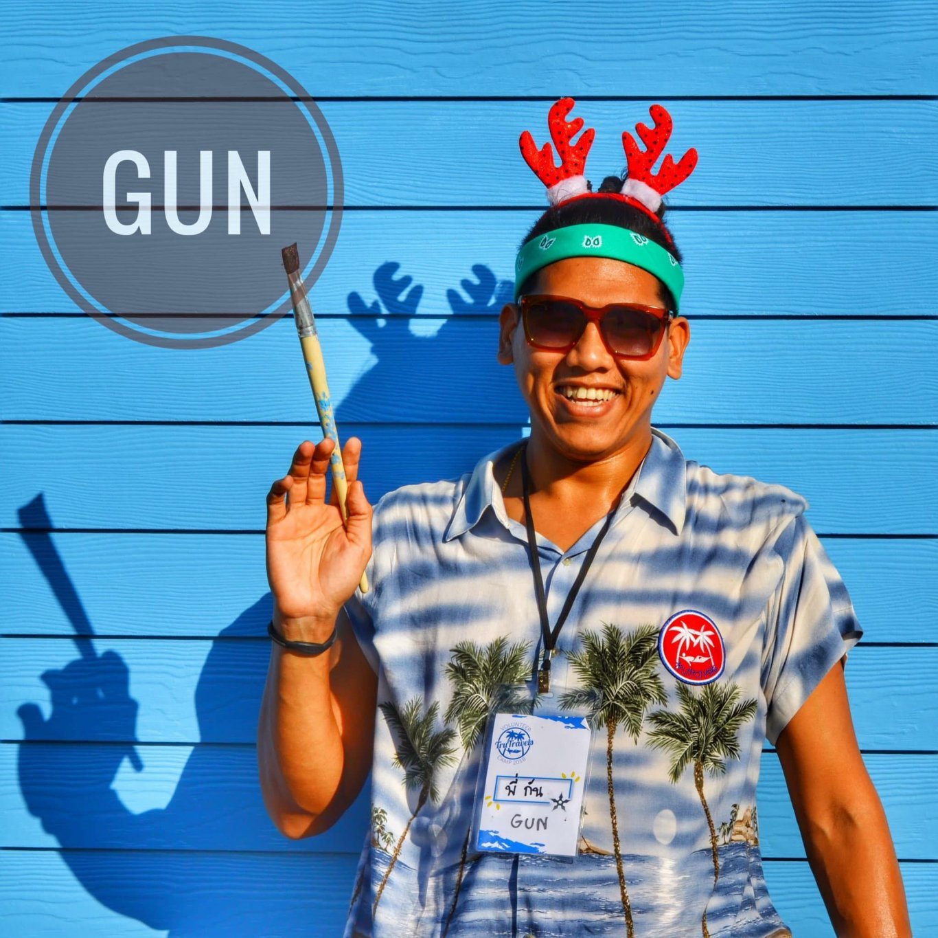Man wearing colourful shirt wearing red Christmas antlers holding paint brush stood in front of bright blue backdrop