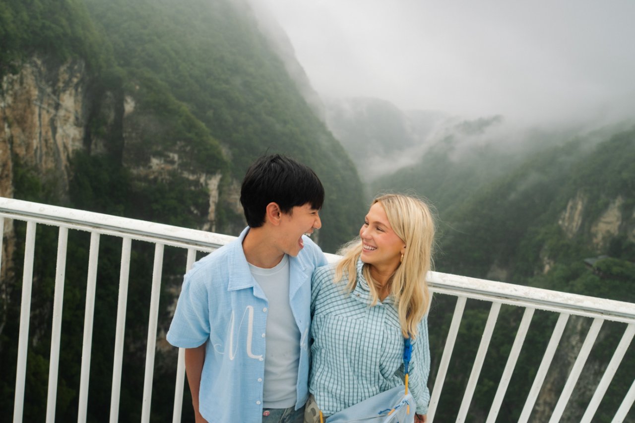 Two travellers laughing together on a bridge above a mist-covered gorge in Zhangjiajie