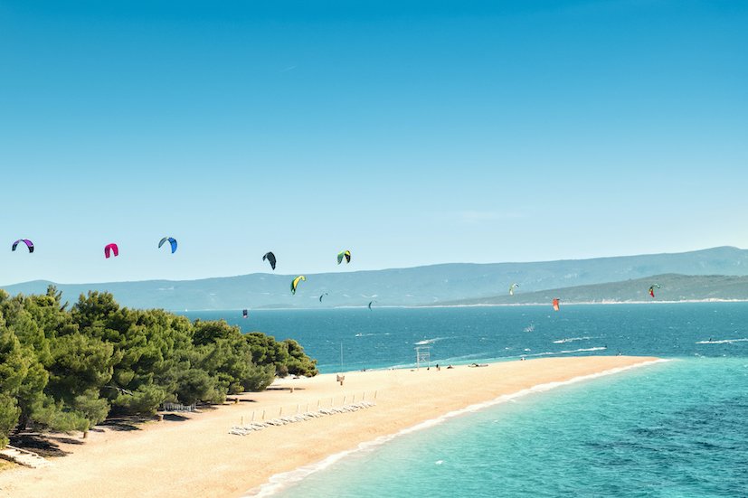 A beach on the island of Brac, Croatia with bright clear blue water and white sandy beach with kite surfing in the background