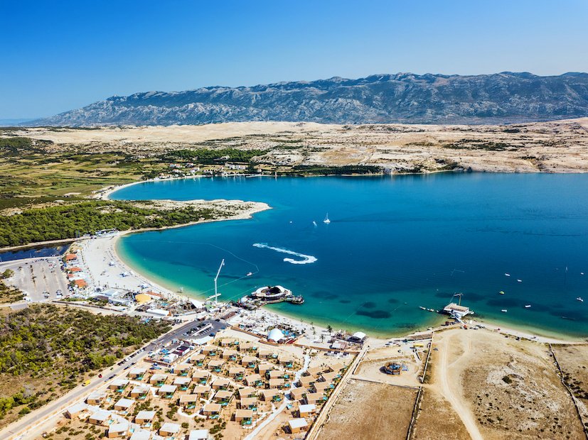 An aerial shot of Pag island in Croatia, showing mountains in the distance, the different shades of blue in the sea, and the town/harbour.
