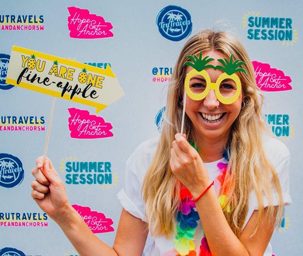 Girl holding pineapple glasses to face holding yellow sign with TruTravels summer party backdrop 