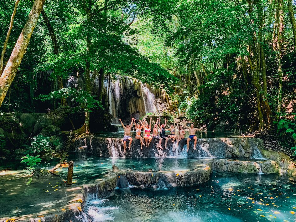 A group sat at the edge of the waterfall on Moyo Island, in Indonesia showing the serene blue waterfall and lush surrounding greenery