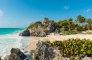 A scenic shot of the Ancient ruins on the beach in Tulum, Mexico showing the pristine sand, bright blue ocean and sky with luscious surrounding greenery & palm trees