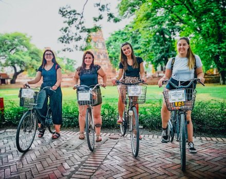 Four girls sitting on bikes with basket on the front with temples faintly showing in the background and green trees behind them