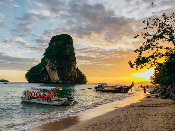 A scenic shot of Railay beach, Krabi, Thailand at sunset with long tail boats and a cliff formation in view